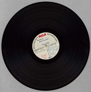 A vinyl record sits against a gray background. On the paper in its center is David Bowie's signature in blue ink.