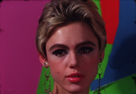 A film still of a person's face in front of a colorful background. The person has short, blonde hair and is wearing makeup and earrings.