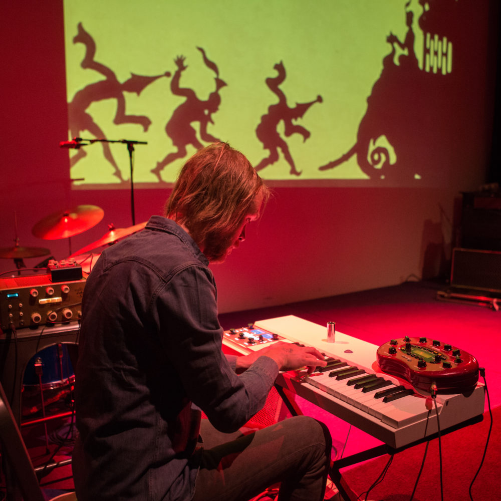 A man with shoulder-length blond hair sits at a keyboard on a stage bathed in red light. In the background, silhouettes of figures wearing hats and an elephant are projected against a yellow-green background.