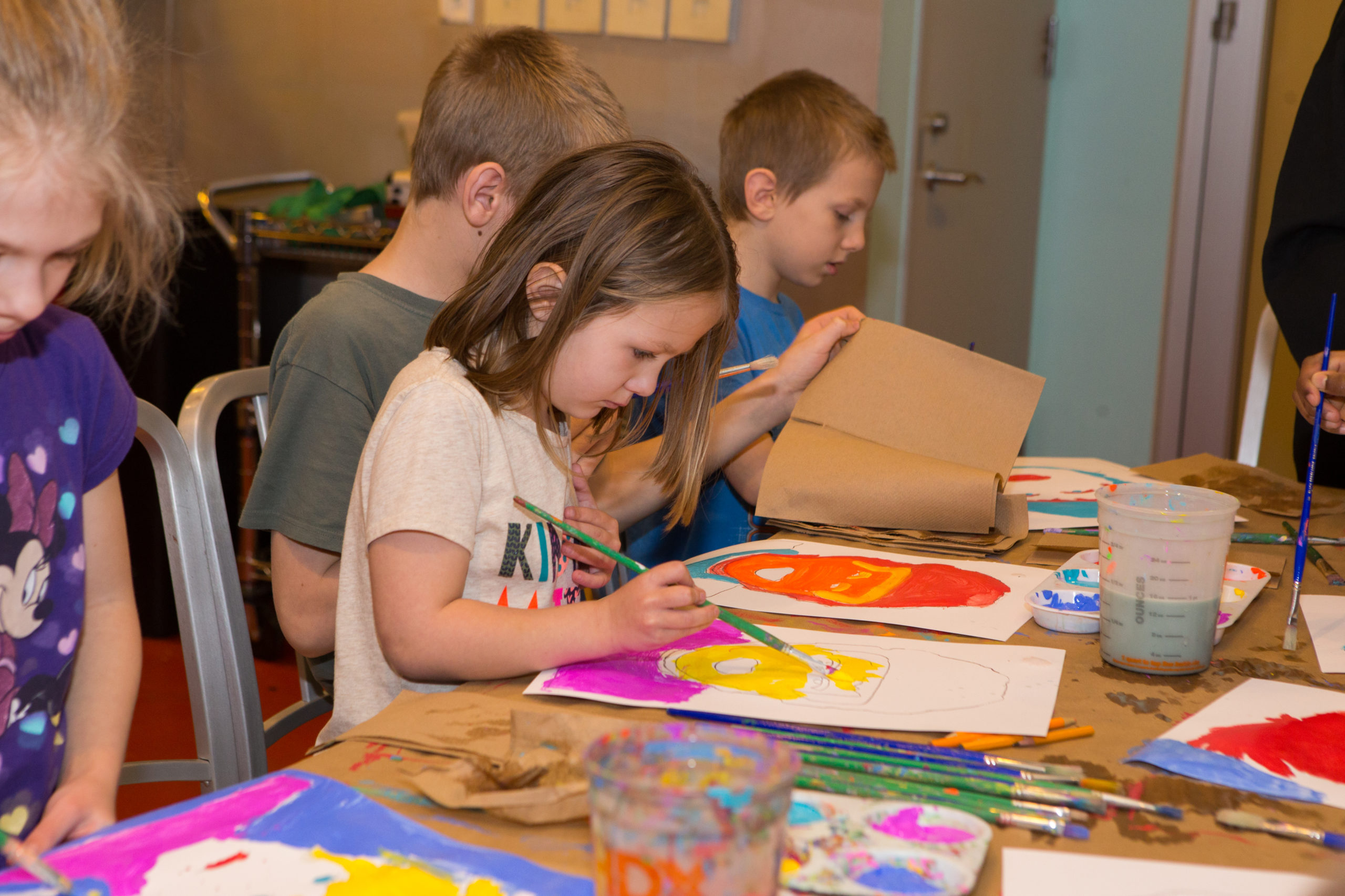 Four children are seated at a table painting colorful portraits.