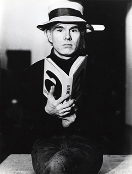 A black and white photograph of Andy Warhol. Warhol poses wearing a white hat with a dark ribbon tied around it and a long-sleeved black shirt. He holds an open book in front of his chest.