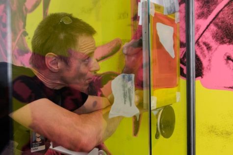 Through a glass door, we see a man with short brown hair who looks very focused as he attaches a canvas to the glass. The wall behind him is covered in bright yellow wallpaper with large pink cows.